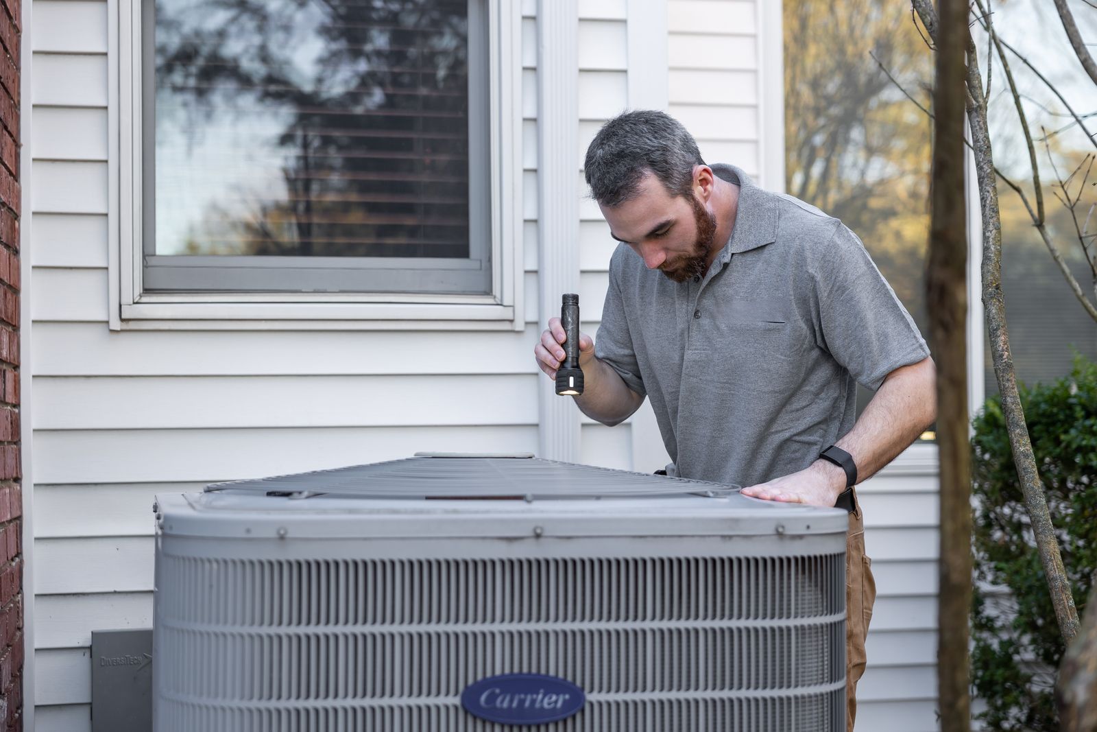 HVAC technician inspecting an outdoor air conditioning unit with a flashlight during a maintenance visit
